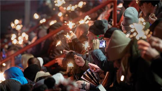 Menschenmenge auf Tribüne hält Wunderkerzen, eine Person filmt mit Smartphone, mehrere tragen Mützen und Jacken.