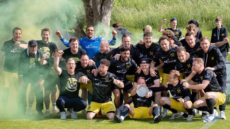 Gruppenfoto einer Fußballmannschaft mit Spielern in schwarzen Trikots und gelben Hosen, die auf einer Wiese jubeln und eine Trophäe halten