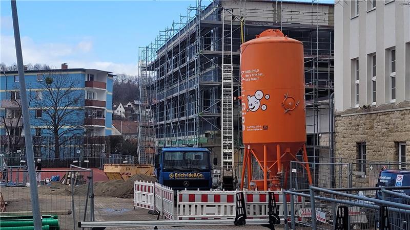 Baustelle mit orangefarbenem Zylinderbehälter, Baugerüst an einem mehrstöckigen Gebäude und mehreren Fahrzeugen im Hintergrund