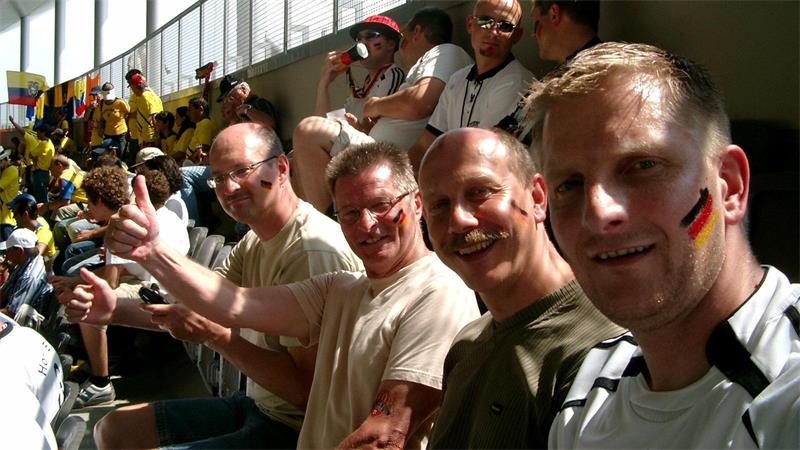 Gruppe von Menschen sitzt auf Tribünenplätzen, einige zeigen Daumen hoch, im Hintergrund weitere Zuschauer in gelben T-Shirts.