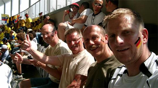 Gruppe von Menschen sitzt auf Tribünenplätzen, einige zeigen Daumen hoch, im Hintergrund weitere Zuschauer in gelben T-Shirts.