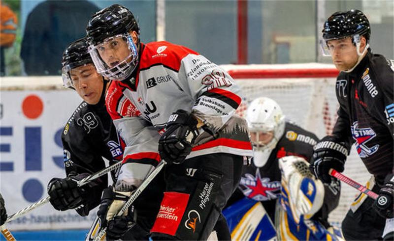 Eishockeyspieler in Aktion auf dem Eis, zwei Spieler in weißen Trikots und zwei in schwarzen Trikots kämpfen um den Puck vor dem Torwart in gelb-blauer Ausrüstung.