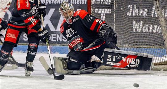 Eishockeyszene vor dem Tor, Torwart in schwarzer Ausrüstung hält den Puck mit dem Schläger.