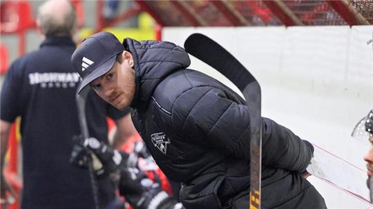 Person in schwarzer Winterjacke lehnt sich an die Bande einer Eishockeybank, im Vordergrund Eishockeyschläger