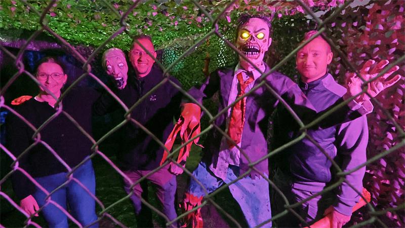 Drei Personen stehen hinter einem Maschendrahtzaun neben zwei Halloween-Figuren mit leuchtenden Augen in einem Garten bei Nacht
