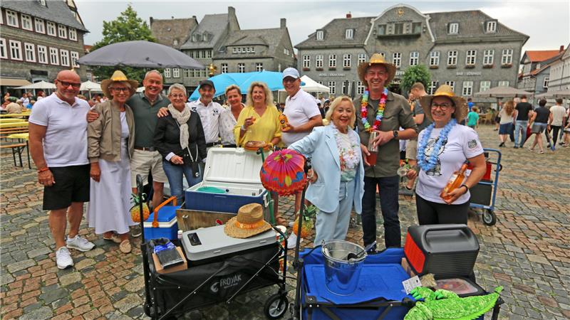 Gruppe von Menschen steht auf einem gepflasterten Platz mit Tischen, Stühlen und Kühlboxen, einige tragen bunte Accessoires wie Hüte und Blumenketten