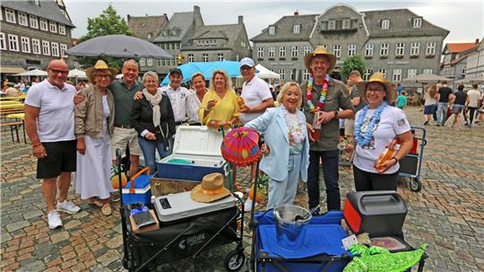Gruppe von Menschen steht auf einem gepflasterten Platz mit Tischen, Stühlen und Kühlboxen, einige tragen bunte Accessoires wie Hüte und Blumenketten