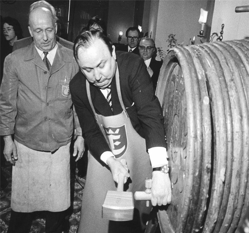 Person mit Schürze schlägt mit Holzhammer auf einen großen Holzfasshahn ein, weitere Personen im Hintergrund