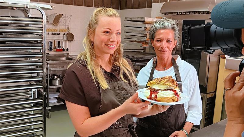 Zwei Frauen in Schürzen in einer Bäckerei, eine hält einen Teller mit einem Stück Kuchen und beide lächeln in eine Kamera