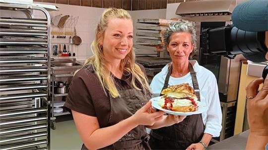 Zwei Frauen in Schürzen in einer Bäckerei, eine hält einen Teller mit einem Stück Kuchen und beide lächeln in eine Kamera