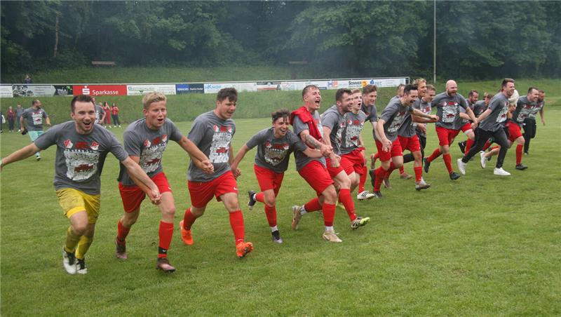 Gruppe von Fußballspielern in grauen Trikots und roten Hosen hält sich an den Händen und springt auf einem Spielfeld