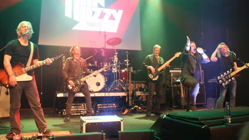 Sechs Musiker auf einer Bühne mit Gitarren, Schlagzeug und Mikrofonen, im Hintergrund ein beleuchtetes Logo mit dem Schriftzug 'LIZZY TRIBUTE'.