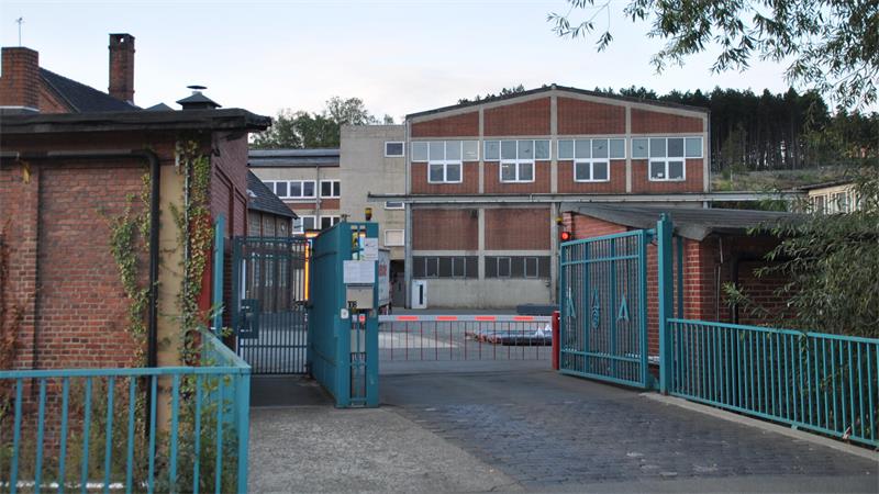 Eingangstor mit blauer Metallumzäunung vor einem mehrstöckigen Backsteingebäude mit großen Fenstern