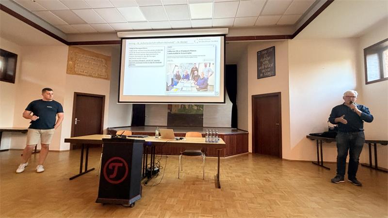 Zwei Männer stehen in einem hellen Raum mit Holzboden vor einem Tisch und einer Leinwand mit einer Präsentation, die Text und eine Illustration zeigt.