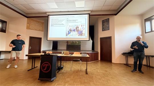 Zwei Männer stehen in einem hellen Raum mit Holzboden vor einem Tisch und einer Leinwand mit einer Präsentation, die Text und eine Illustration zeigt.