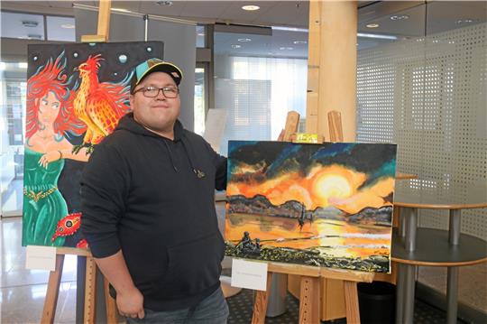 Florian Wallrich stellt seine Kunstwerke in der Volksbank aus. „Der Sonnenuntergang“ ist eines seiner Lieblingsbilder. Wer genau hinschaut, entdeckt klein im Vordergrund den Künstler selbst und seinen Dackel-Jack-Russel-Mischling Rocky. Links im Bild „Das Erwachen des Phönix“. Eigentlich hatte er nur den Feuervogel allein malen wollen, doch der Künstler merkt bald: „Da fehlt was.“ So kam noch eine Frau ins Bild, die den Phönix beschwören kann.  Fotos: Hartmann