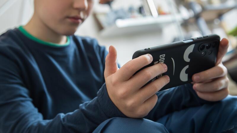 Kind sitzt mit verschränkten Beinen und hält ein Smartphone mit schwarzer Hülle in beiden Händen