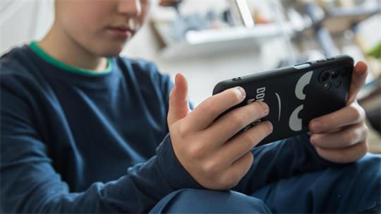 Kind sitzt mit verschränkten Beinen und hält ein Smartphone mit schwarzer Hülle in beiden Händen