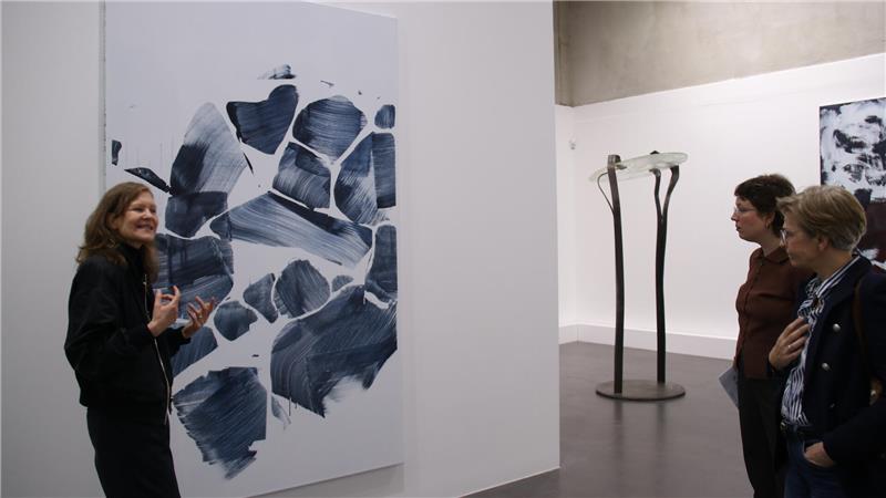 Drei Personen betrachten abstraktes schwarz-weißes Gemälde in einer Galerie mit weiterer Skulptur im Hintergrund.
