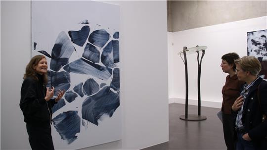 Drei Personen betrachten abstraktes schwarz-weißes Gemälde in einer Galerie mit weiterer Skulptur im Hintergrund.