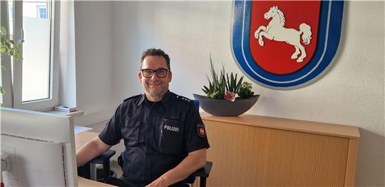 Polizist in Uniform sitzt an einem Schreibtisch in einem Büro mit einem Wappen an der Wand, das ein springendes Pferd zeigt.