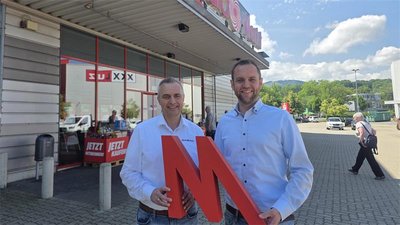 Zwei Männer in weißen Hemden stehen vor einem Gebäude mit dem Schriftzug 'Media Markt' und halten gemeinsam ein großes rotes 'M'.