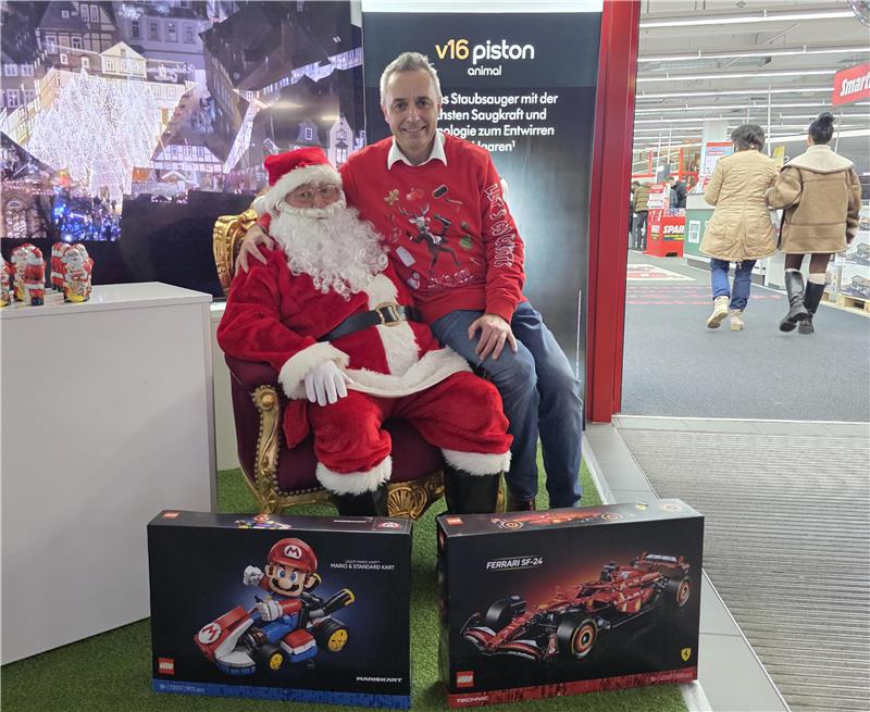 Weihnachtsmann in rotem Kostüm sitzt auf einem goldverzierten Stuhl, daneben Person mit rotem Pullover, davor zwei Spielzeugboxen mit Rennwagen und Spielfigur.