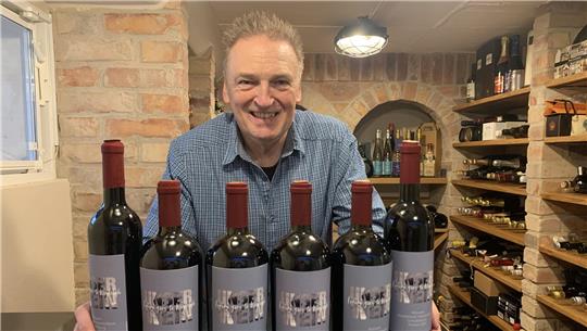 Mann in blau kariertem Hemd hält sechs Flaschen Wein mit roten Kapseln in einem Weinkeller mit Backsteinwänden und Holzregalen