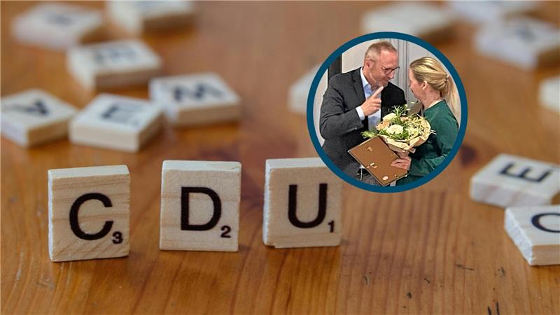 Ein Mann im Anzug zeigt mit dem Zeigefinger auf eine Frau mit blondem Pferdeschwanz, die einen Blumenstrauß hält.