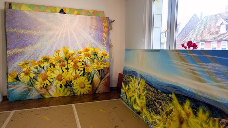 Fred Hahns Werke im Atelier: Gelbe Blumen im Sonnenlicht, was sich in verschiedene Farben aufspaltet, und eine bergige Landschaft mit Blumen und Insekten.