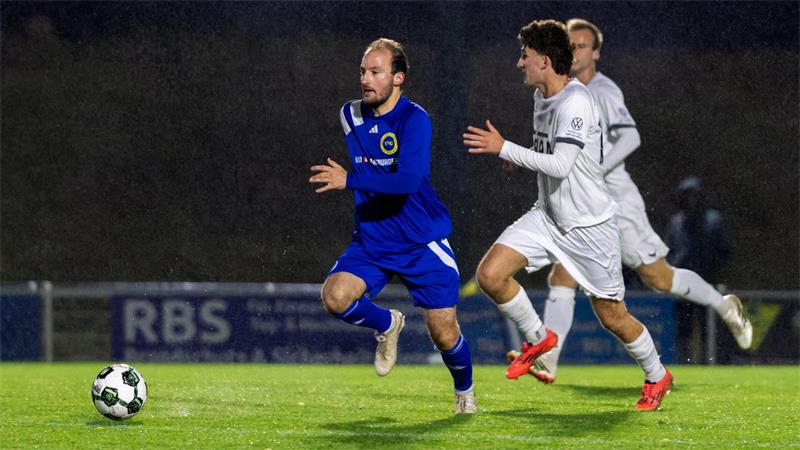 Ein Spieler in blauer Kleidung und zwei Spieler in weißer Kleidung laufen einem Fußball hinterher