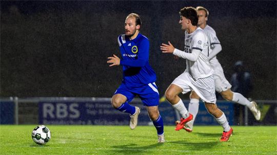 Ein Spieler in blauer Kleidung und zwei Spieler in weißer Kleidung laufen einem Fußball hinterher