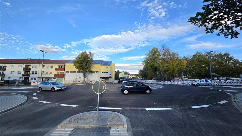 Kreisverkehr mit neu asphaltierter Fahrbahn und Verkehrsinsel in einer Wohnsiedlung mit mehrstöckigen Wohnhäusern und Bäumen im Hintergrund