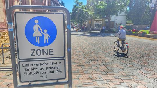 Verkehrsschild mit Symbol einer Frau und einem Kind, darunter Text zu Lieferverkehr und Zufahrt, im Hintergrund eine gepflasterte Straße mit einem Radfahrer.