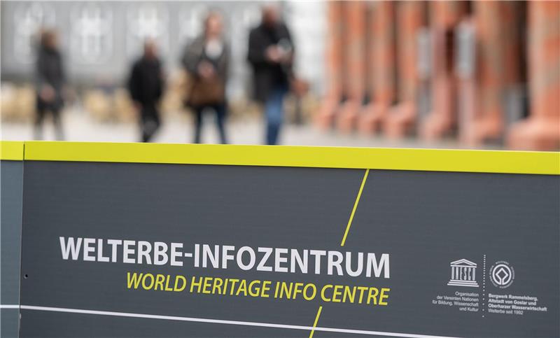 Schild mit der Aufschrift 'WELTERBE-INFOZENTRUM WORLD HERITAGE INFO CENTRE' vor unscharf erkennbaren Personen und Gebäuden im Hintergrund