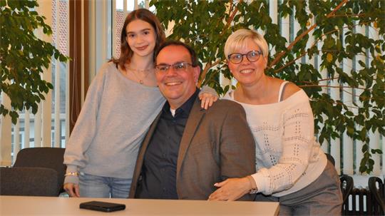 Drei Personen lächeln sitzend und stehend an einem Tisch vor großen Fenstern mit grünen Pflanzen