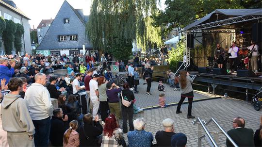 Zwischen Häusern und Bühne tummeln sich viele Zuschauer, ein paar tanzen vor der Bühne, auff der eine Band spielt.