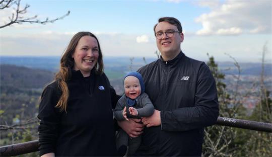 Drei Personen stehen vor einer Landschaft mit Hügeln und Bäumen, zwei Erwachsene halten ein lächelndes Baby