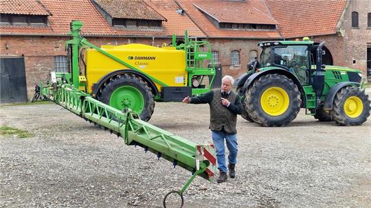 Mann steht vor großem grünen und gelben Traktor mit angebautem landwirtschaftlichen Gerät auf gepflastertem Hof vor Backsteingebäude