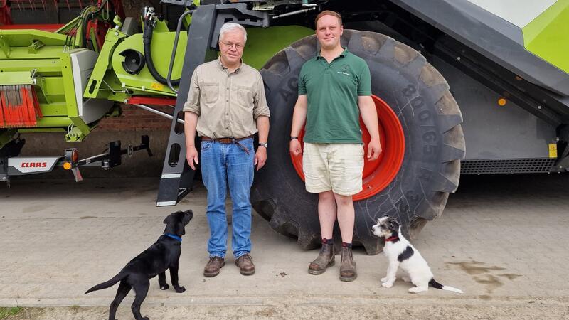 Zwei Männer stehen vor einem großen landwirtschaftlichen Fahrzeug mit zwei Hunden auf dem Boden