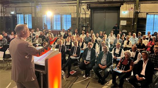 Viele Menschen sitzen nebeneinander, einer spricht auf dem Podium.