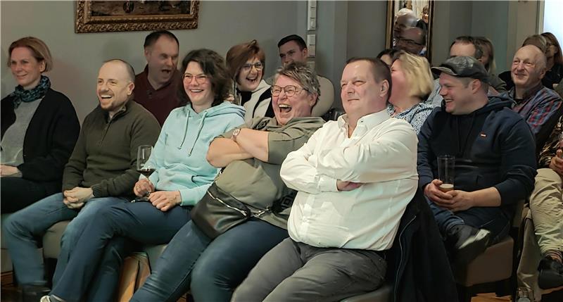 Eine fröhliche Gruppe von Menschen sitzt in einem Raum auf Stühlen, einige mit verschränkten Armen,.
