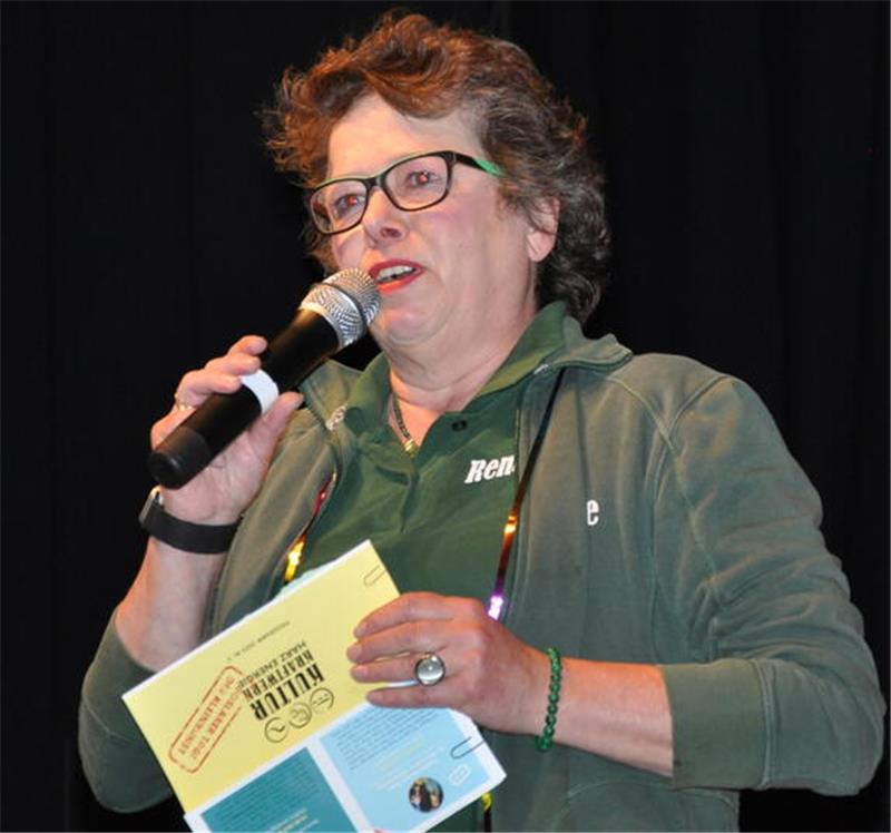 Frau mit lockigem Haar hält ein Mikrofon in der rechten Hand und ein aufgeschlagenes Programmheft in der linken Hand, trägt ein grünes Oberteil und eine grüne Jacke.