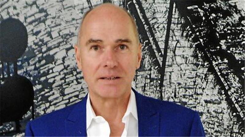 Ein Mann mit Glatze trägt ein blaues Sakko und ein weißes Hemd vor einer Wand mit schwarzer und weißer abstrakter Musterung.