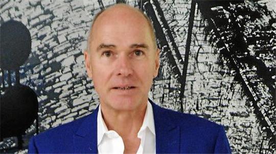 Ein Mann mit Glatze trägt ein blaues Sakko und ein weißes Hemd vor einer Wand mit schwarzer und weißer abstrakter Musterung.