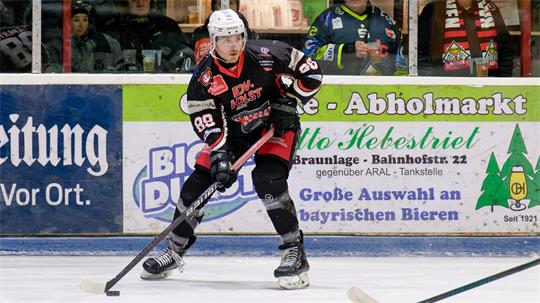 Eishockeyspieler in schwarzer Ausrüstung mit weißem Helm steuert den Puck auf dem Eis, im Hintergrund Zuschauer hinter der Bande.
