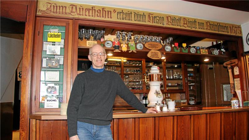 Mann lehnt an einer Holztheke in einem Gastraum mit Gläsern, Bierkrügen und einem großen Zapfhahn hinter der Theke.