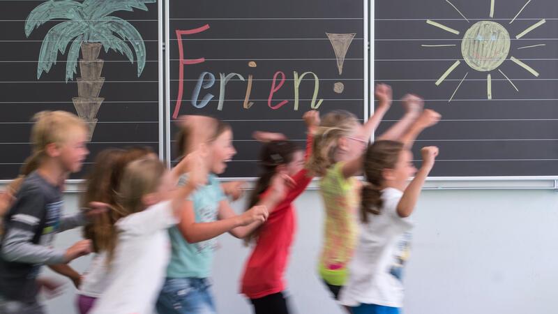 Kinder laufen in einem Klassenzimmer vor einer Tafel mit der Aufschrift "Ferien!" vorbei.