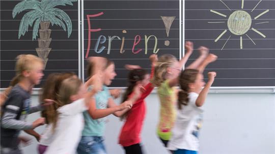 Kinder laufen in einem Klassenzimmer vor einer Tafel mit der Aufschrift "Ferien!" vorbei.