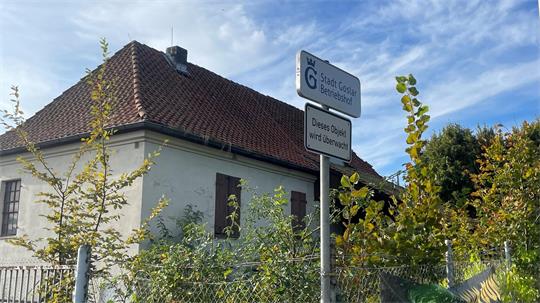 Haus mit rotem Ziegeldach und zwei Verkehrsschildern vor grünem Buschwerk unter blauem Himmel